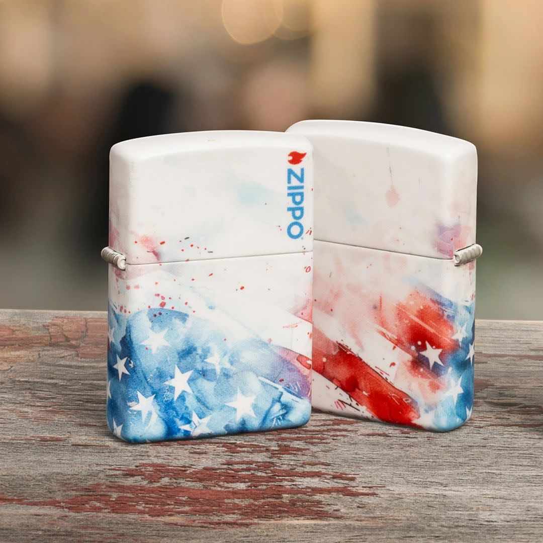 Lifestyle image of two Zippo Watercolor Flag Design 540 Matte Windproof Lighters both standing on a weathered wooden surface; one showing the front and hinge side of the lighter while the other is showing the back and hinge side of the lighter. 

