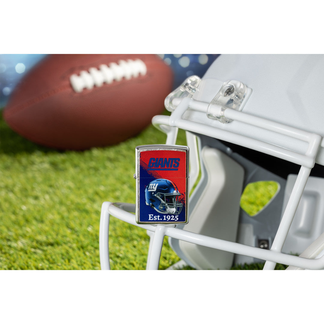 Lifestyle image of Zippo NFL New York Giants Helmet Design Street Chrome Windproof Lighter standing on the facemask of a white helmet showing the front of the lighter with a football lying on a football field as the background.

