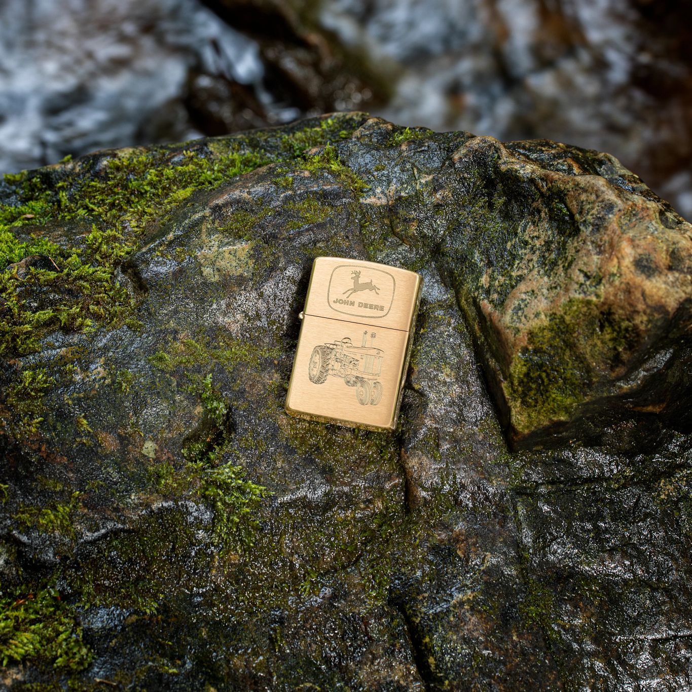 Lifestyle image of Zippo John Deere Tractor Design Brushed Brass Windproof Lighter lying on a mossy boulder with another boulder in the background. 

