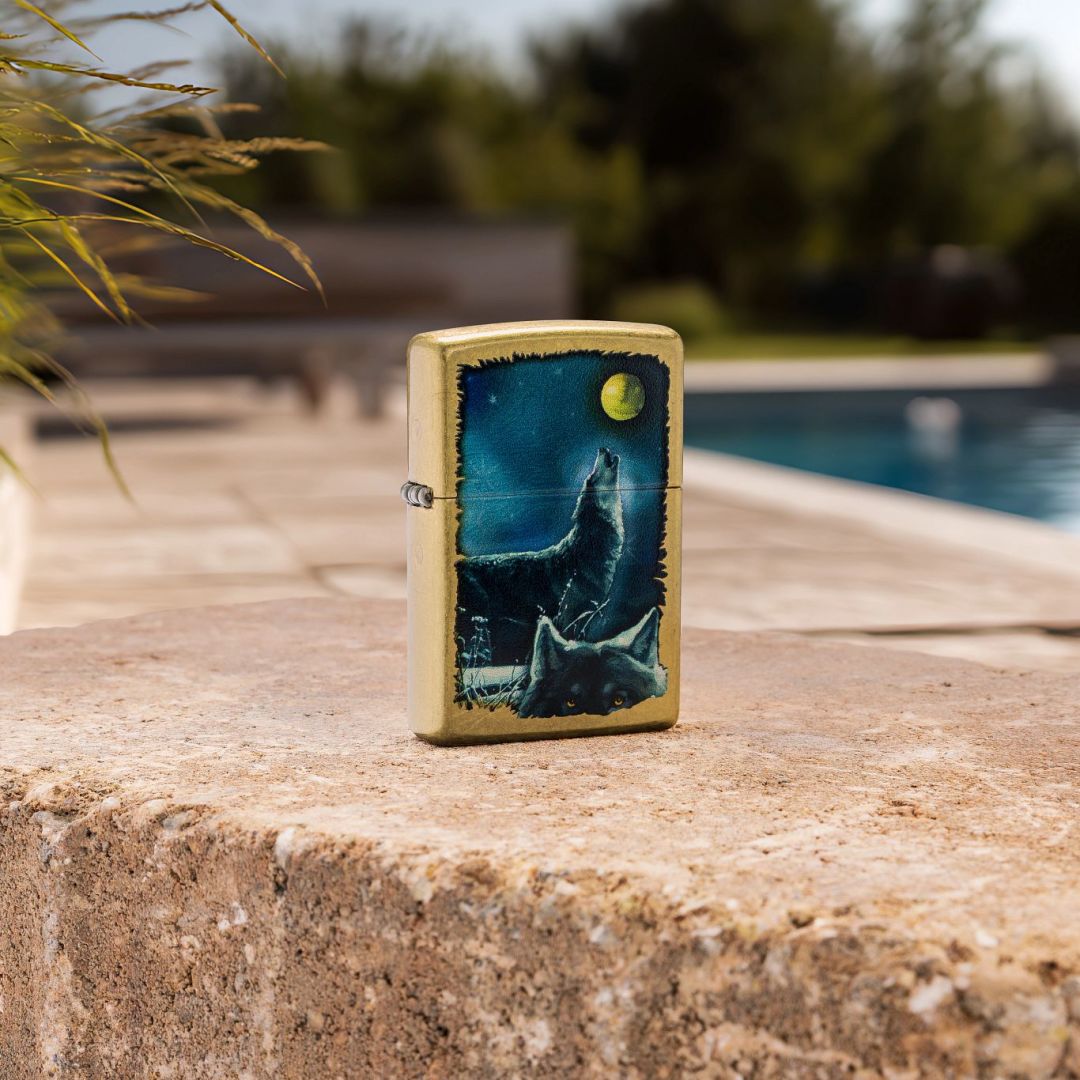Lifestyle image of Zippo Wolf Song Design Street Brass Windproof Lighter standing next to an outdoor pool while showing the front and hinge side of the lighter with the pool and trees as the background. 

