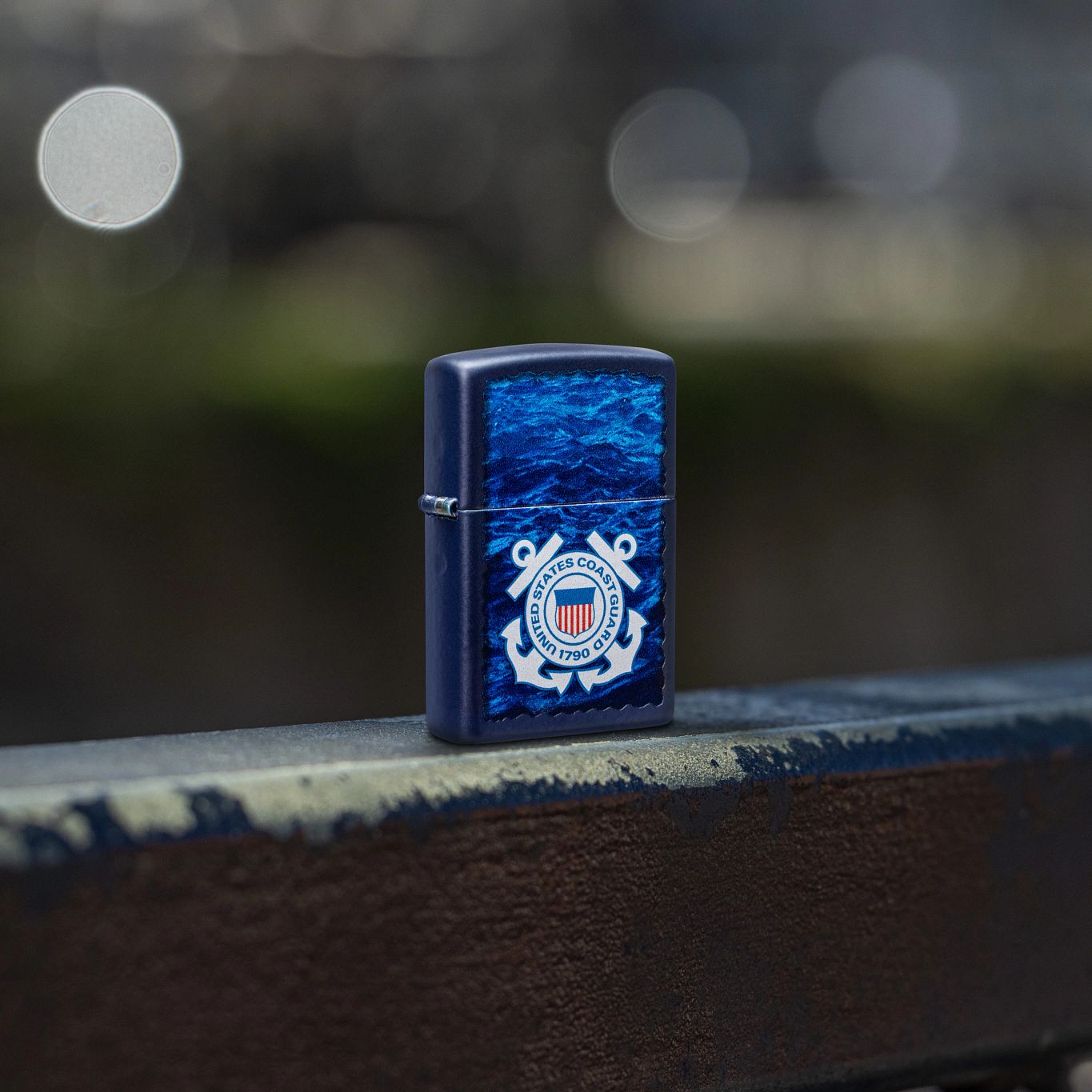 Lifestyle image of Zippo U.S. Coast Guard Waves Design Navy Matte Windproof Lighter standing on a dark painted railing showing the front and hinge side of the lighter. 

