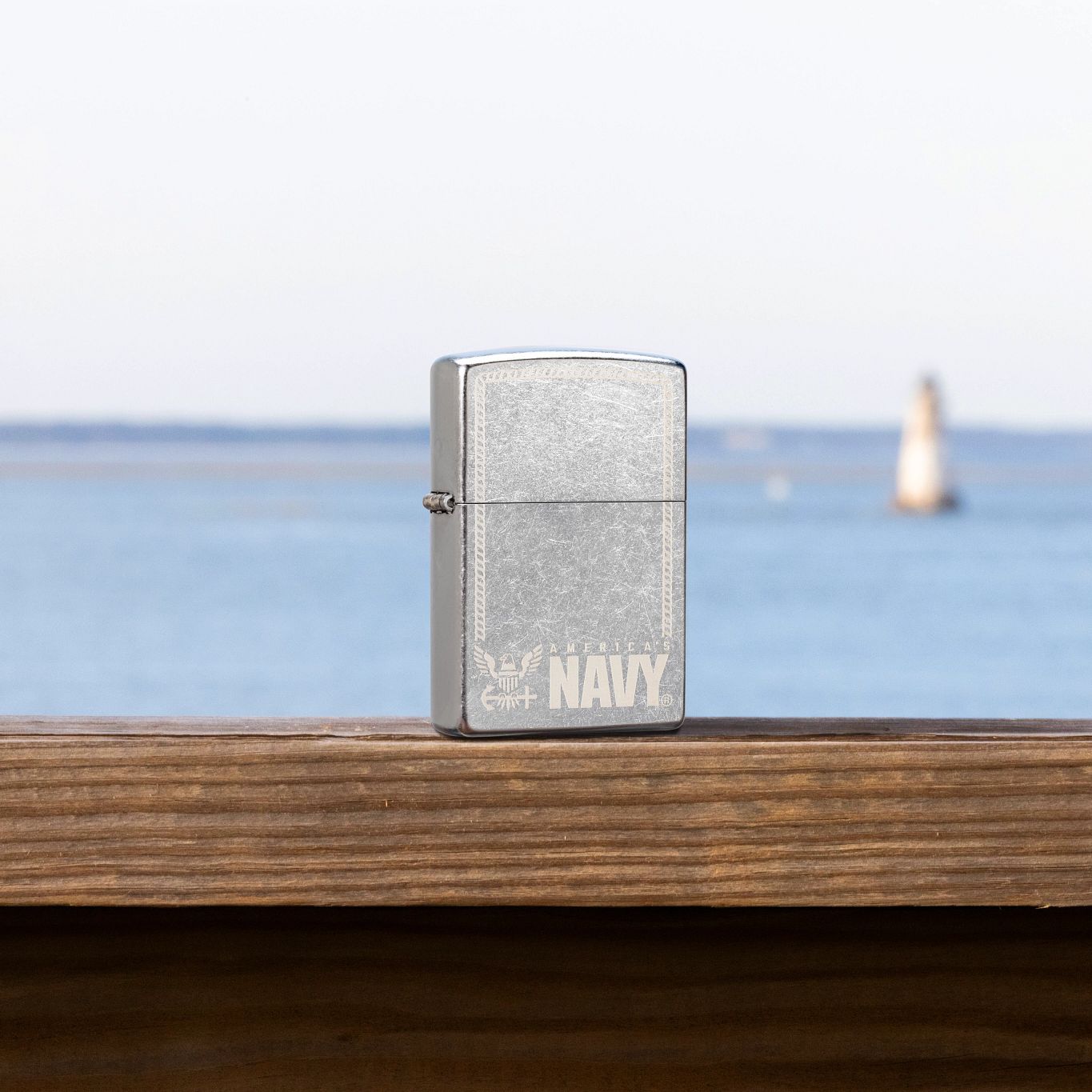 Lifestyle image of Zippo United States Navy® America's Navy Design Street Chrome Windproof Lighter standing on a wooden railing with a body of water as the background, showing the front and hinge side of the lighter.