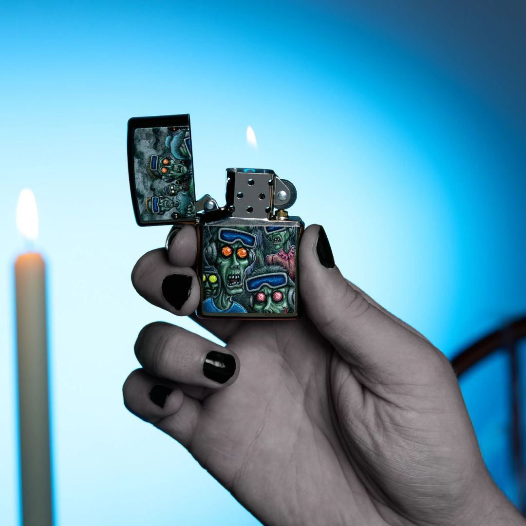 Lifestyle image of Zippo Zombie Escape Design Black Ice® Windproof Lighter open and lit in hand showing the front of the lighter with a lit white taper candle and a blue ombre background.

