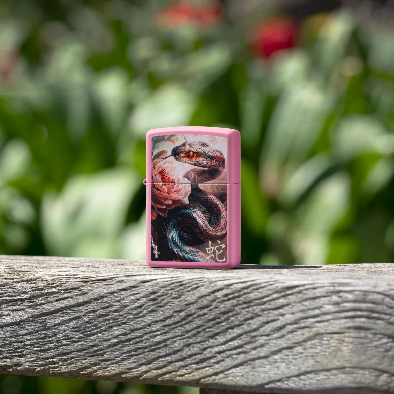 Lifestyle image of Zippo Mazzi® Snake and Flower Design Pink Matte Windproof Lighter standing on a wooden bench with blurred flowers behind it.