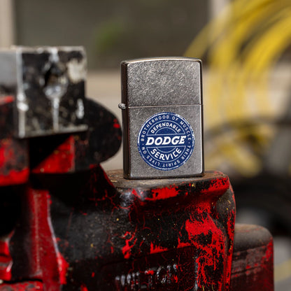 Lifestyle image of Zippo Dodge Service Sign Satin Chrome Windproof Lighter standing on an old mechanics pro vise with a blurred garage in the background. 

