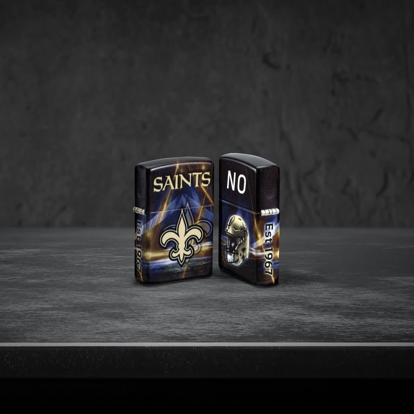 Lifestyle image of two Zippo NFL New Orleans Saints 540 Matte Windproof Lighters, one showing the front of the lighter and the other showing the back, standing on a dark grey surface.