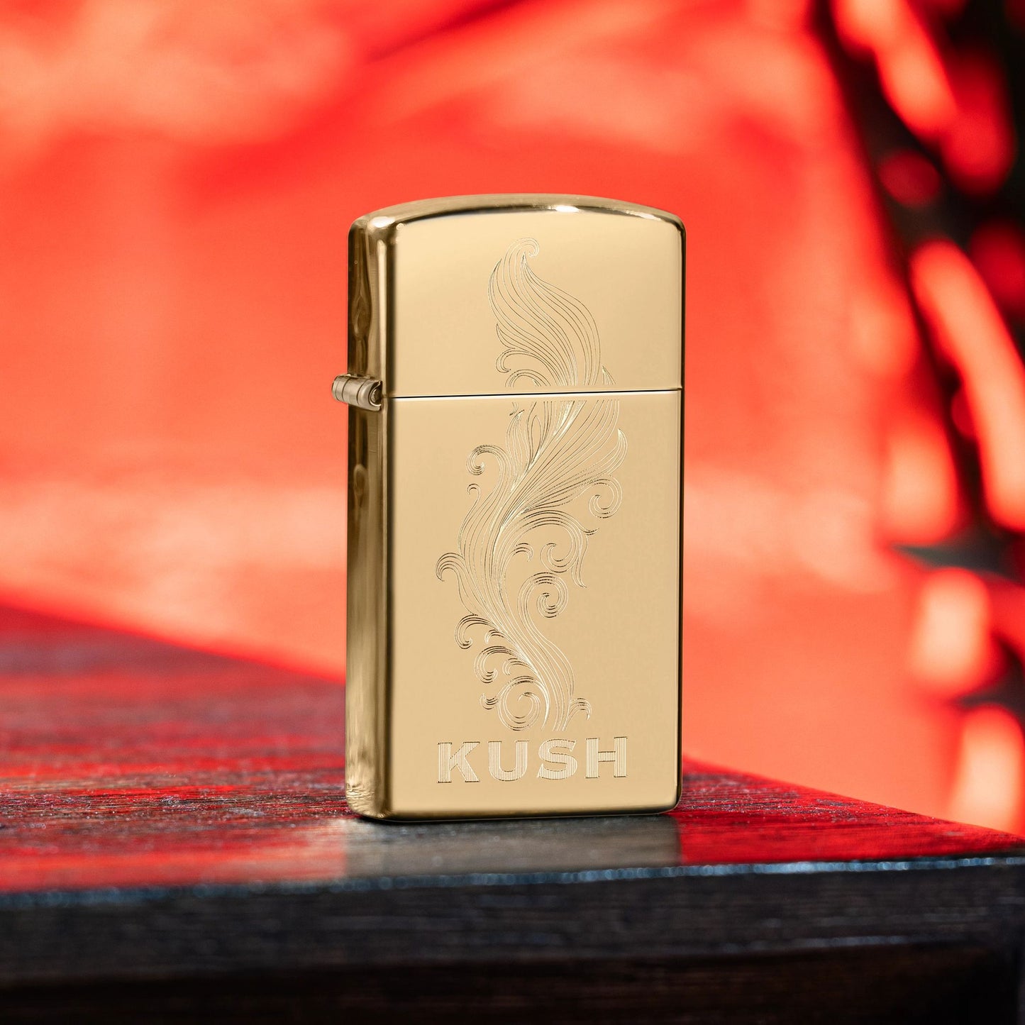 Lifestyle image of Zippo Slim® Kush Design High Polish Brass Windproof Lighter standing on the corner of a dark wooden table showing the front of the lighter with a red background.

