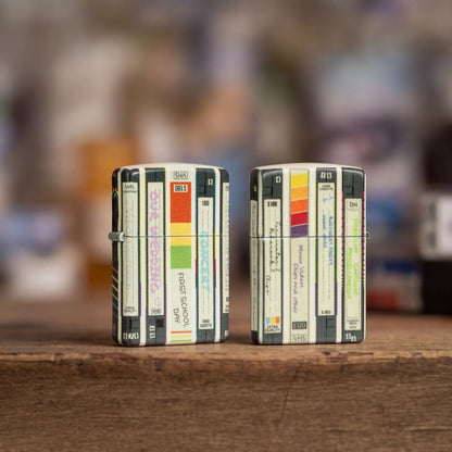 Lifestyle image of two Zippo Home Video Design 540 Matte Windproof Lighters standing on a wooden surface. One showing the front and hinge side of the lighter while the other is showing the back and non-hinge side of the lighter. 

