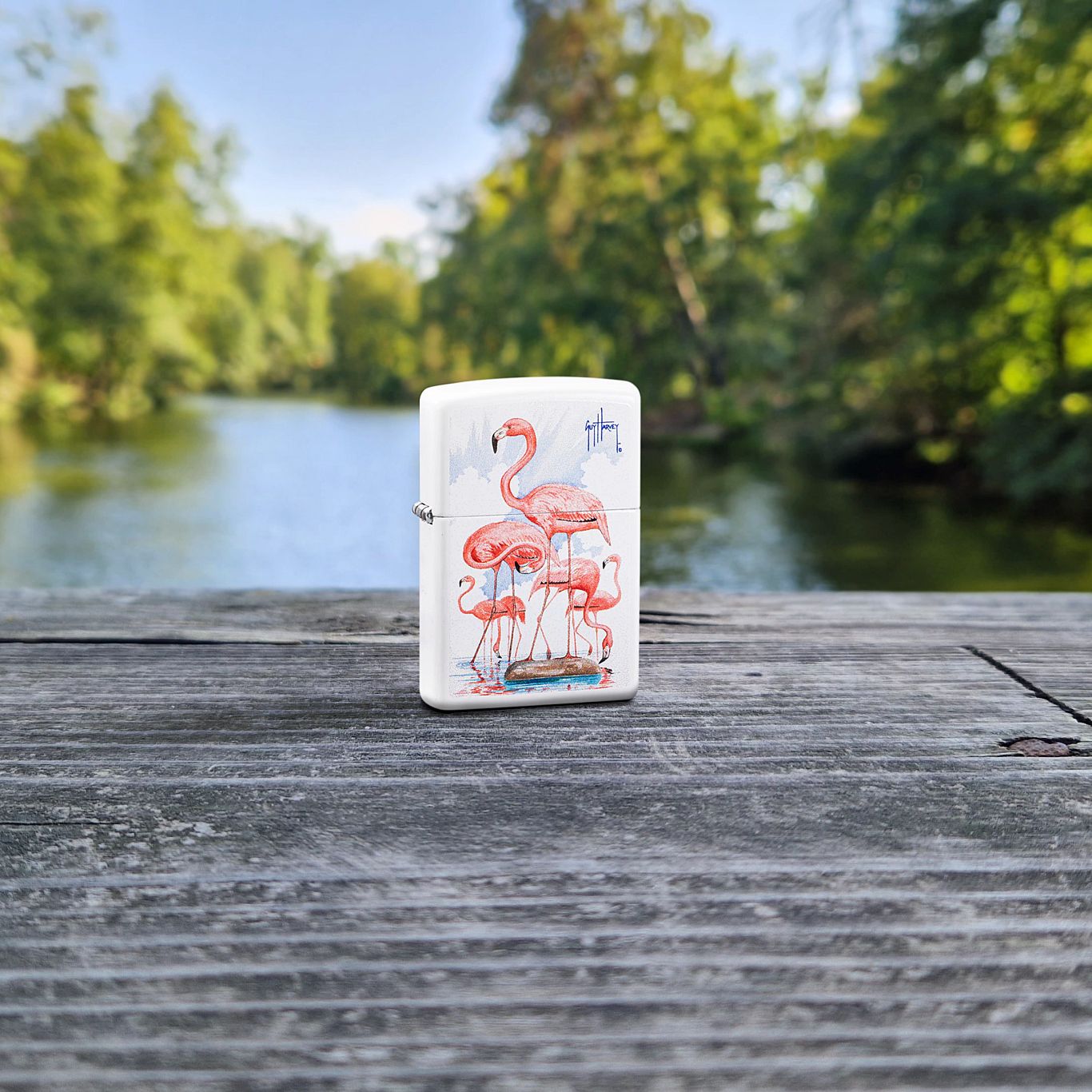 Lifestyle image of Zippo Guy Harvey Flamingos Design White Matte Windproof Lighter standing on a wooden railing with a creek and green trees as the background, showing the front of the lighter. 

