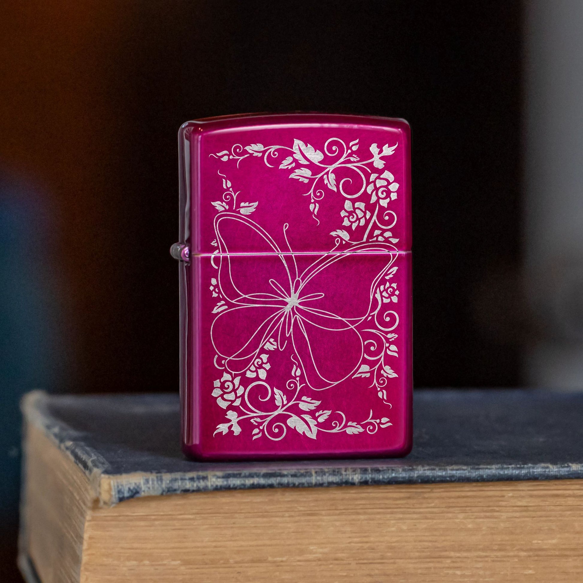 Lifestyle image of Zippo Butterfly Bliss Design Candy Raspberry Windproof Lighter standing on the cover of a book showing the front of the lighter. 

