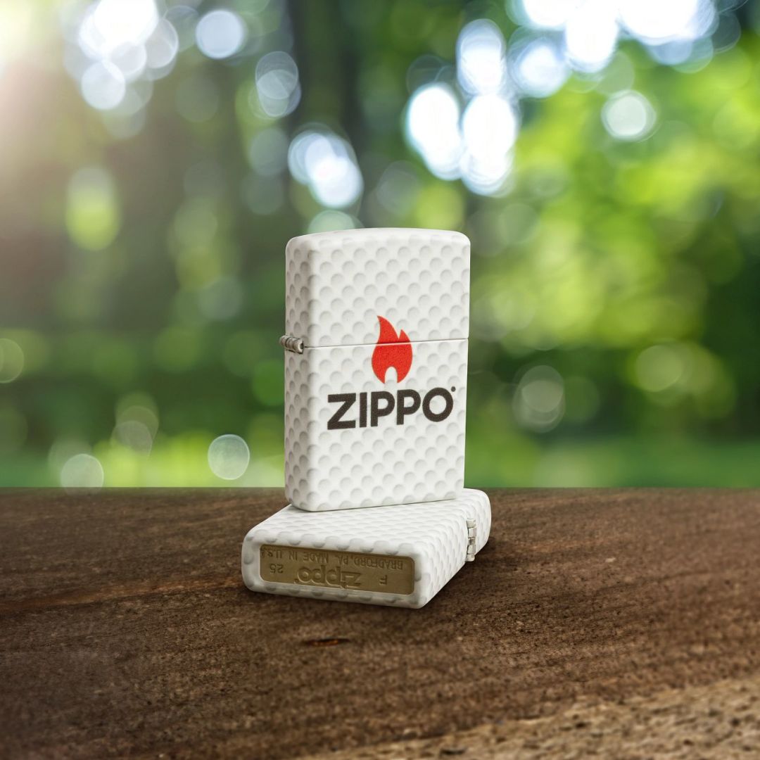 Lifestyle image of two Zippo Golf Ball Design 540 Matte Windproof Lighters; one lying flat on a wooden surface showing the bottom stamp, back and hinge side of the lighter while the other lighter is standing on top of the lighter that is lying flat, showing the front and hinge side of the lighter with an out of focus background with green trees. 

