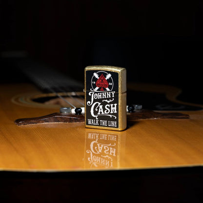 Lifestyle image of Zippo Johnny Cash 1932 Design Street Brass Windproof Lighter standing on the body of an acoustic guitar showing the front and non-hinge side of the lighter with the rest of the guitar as the background. 

