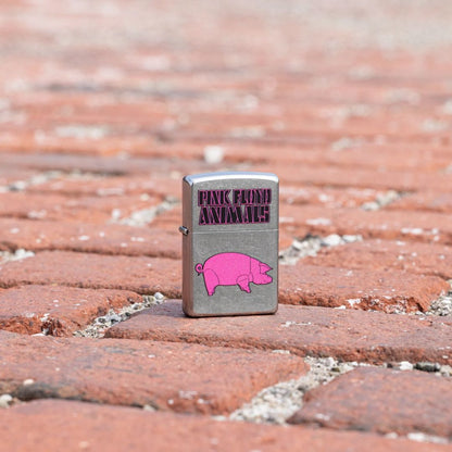 Lifestyle image of Zippo Pink Floyd Animals Design Street Chrome Windproof Lighter standing on a brick walkway showing the front of the lighter with the brick walkway as the background. 

