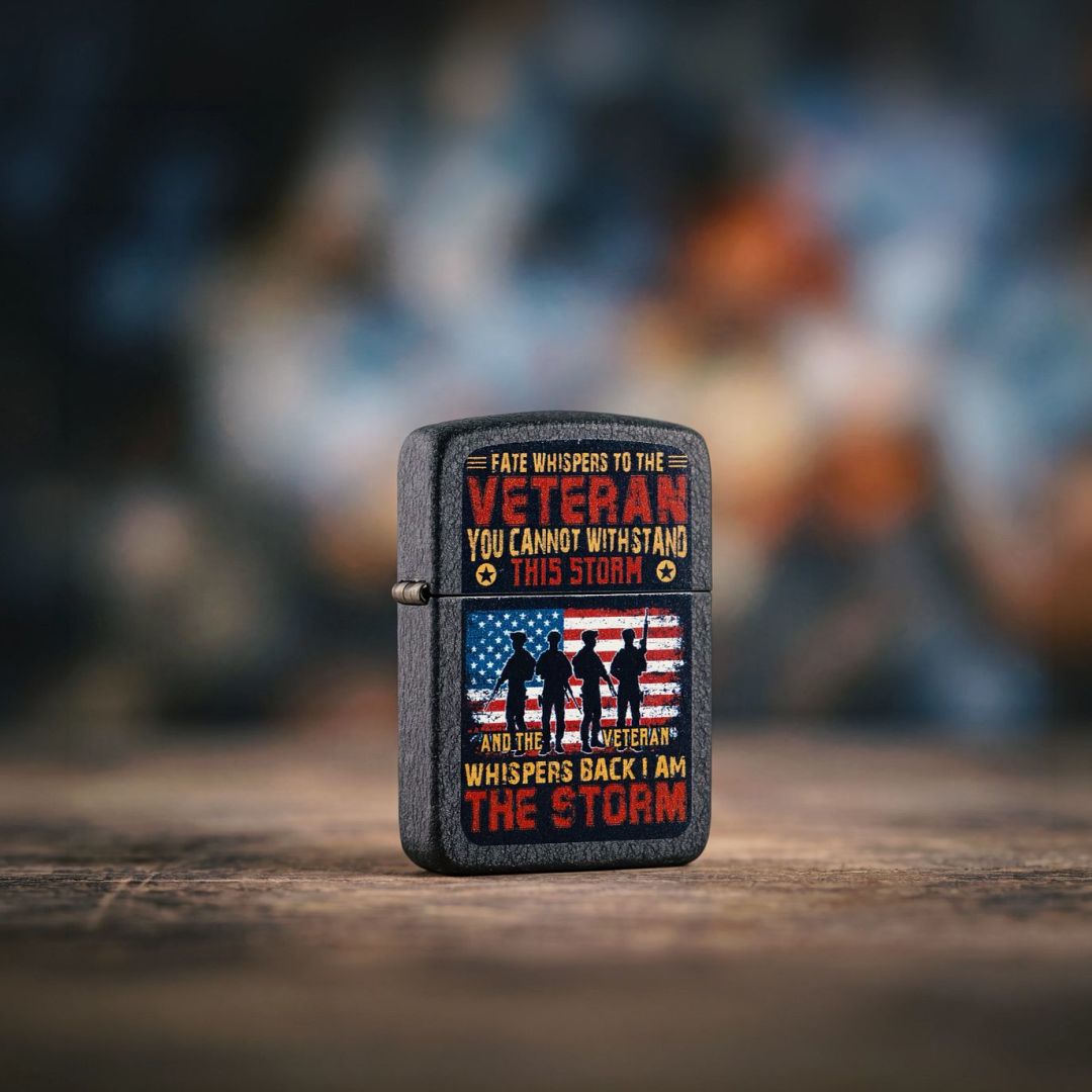 Lifestyle image of Zippo I am the Storm Design 1941 Replica Black Crackle® Windproof Lighter standing on a dark concrete surface showing the front and hinge side of the lighter with a muted out of focus red, white and blue graffiti style background. 

