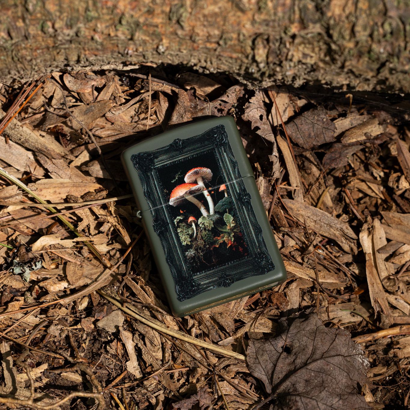 Lifestyle image of Zippo Framed Fungi Design Green Matte Windproof Lighter lying in a mulch and brown fall foliage mixture at the base of a tree trunk.

