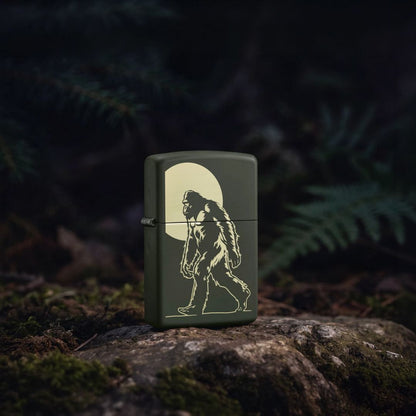 Lifestyle image of Zippo Sasquatch Stroll Design Green Matte Windproof Lighter standing on a rock showing the front and hinge side of the lighter with ferns in a dark background. 

