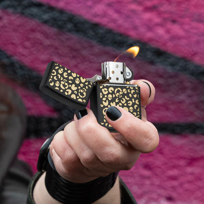 Lifestyle image of Zippo Leopard Skulls Design Black Matte Windproof Lighter being held in hand, open and lit, showing the front of the lighter with a purple and black graffiti background. 

