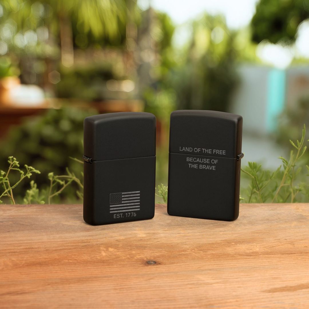 Lifestyle image of two Zippo Because of the Brave Design Black Matte with Chrome Windproof Lighters both standing on a wooden surface; one showing the front and hinge side of the lighter while the other is showing the back of the lighter with an out of focus nature background. 

