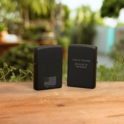 Lifestyle image of two Zippo Because of the Brave Design Black Matte with Chrome Windproof Lighters both standing on a wooden surface; one showing the front and hinge side of the lighter while the other is showing the back of the lighter with an out of focus nature background. 

