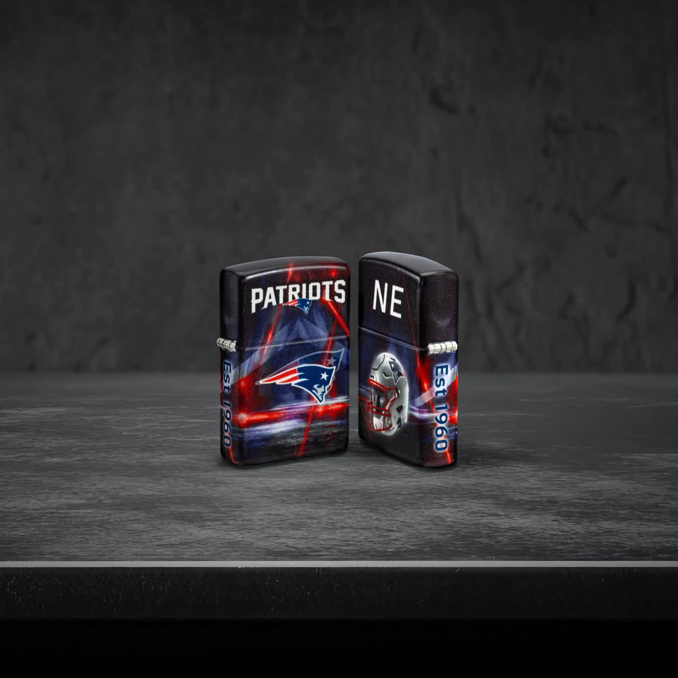 Lifestyle image of two Zippo NFL New England Patriots 540 Matte Windproof Lighters, one showing the front of the lighter and the other showing the back, standing on a dark grey surface.