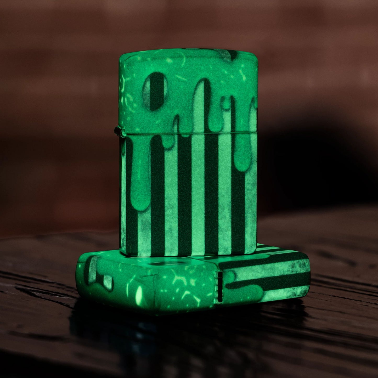 Lifestyle image of two Zippo Glowing Goo Design Glow-in-the-Dark Green Windproof Lighters; one lying flat on a dark wooden table showing the top of the lid and hinge side of the lighter while the other lighter is standing on top of the lighter that is lying flat, showing the front of the lighter while glowing in the dark. 

