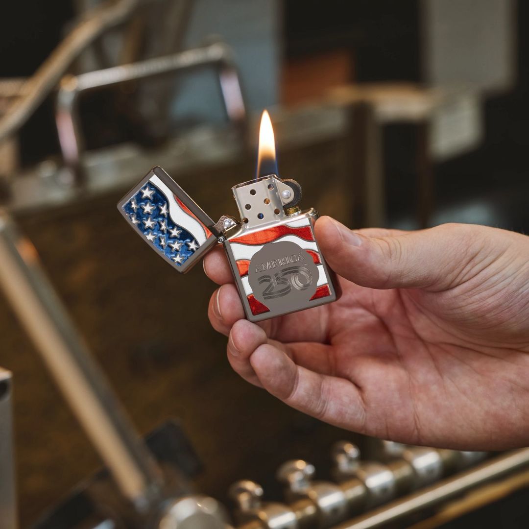 Lifestyle image of Zippo America250 Flag Design Black Ice Windproof Lighter showing the front of the lighter while lit and in hand with a workshop in the background. 

