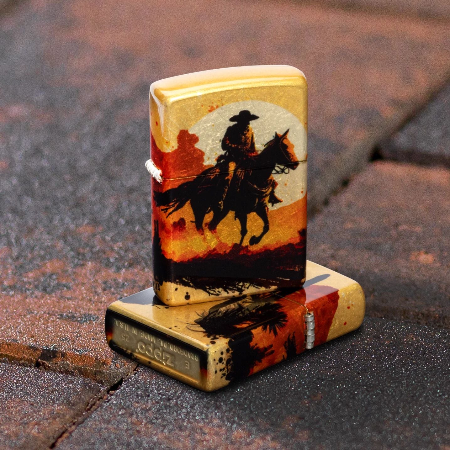 Lifestyle image of two Zippo Racing Wrangler Design 540 Tumbled Brass Windproof Lighters; one lying flat on brick ground showing the bottom stamp and hinge side of the lighter while the other lighter is standing on top of the lighter that is lying flat, showing the front of the lighter. The background is the brick ground. 