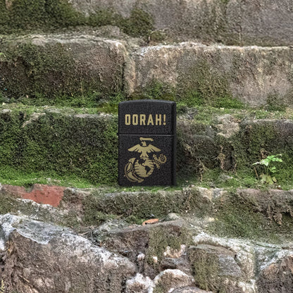 Lifestyle image of Zippo U.S. Marine Corps Oorah Design Black Crackle® Windproof Lighter standing on mossy stone stairs showing the front of the lighter. 

