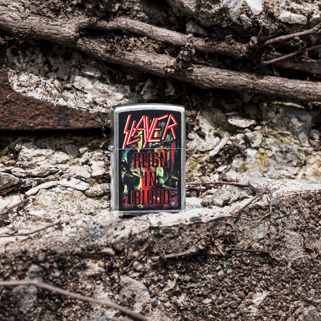 Lifestyle image of Zippo Slayer Reign in Blood Design Street Chrome Windproof Lighter standing on a rocky surface showing the front of the lighter with rocks and twigs in the background. 

