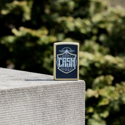Lifestyle image of Zippo Johnny Cash Crest Design Street Brass Windproof Lighter standing on the edge of a concrete block showing the front of the lighter with out of focus trees in the background. 

