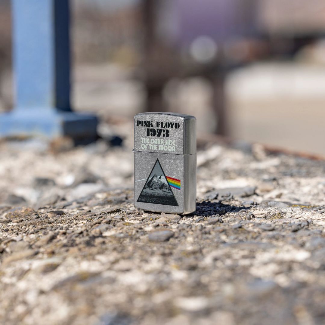 Lifestyle image of Zippo Pink Floyd 1973 Design Street Chrome Windproof Lighter standing on a rocky surface at a 3/4 angle showing the front and non-hinge side of the lighter with an out of focus background. 

