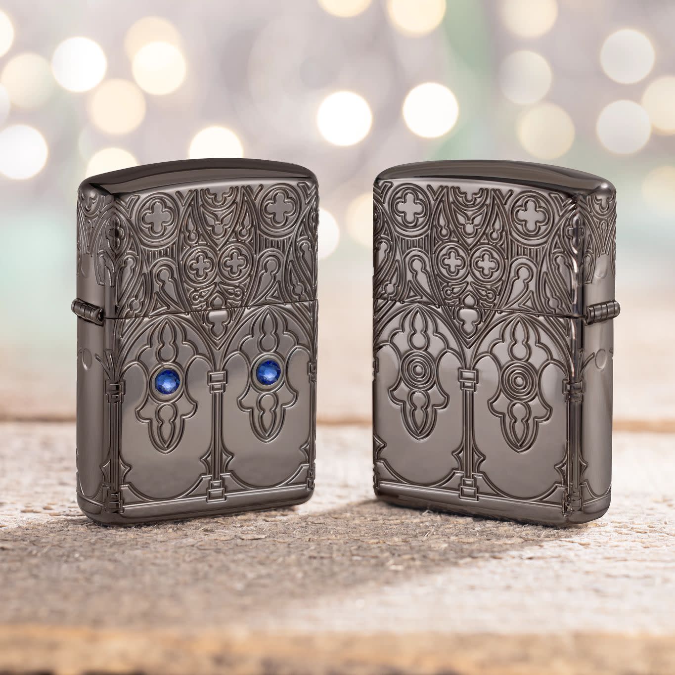 Lifestyle image of two Zippo Gothic Window Design Armor® Black Ice® Windproof Lighters, one showing the front of the lighter and the other showing the back, standing on a cement surface with blurred twinkle lights in the background.