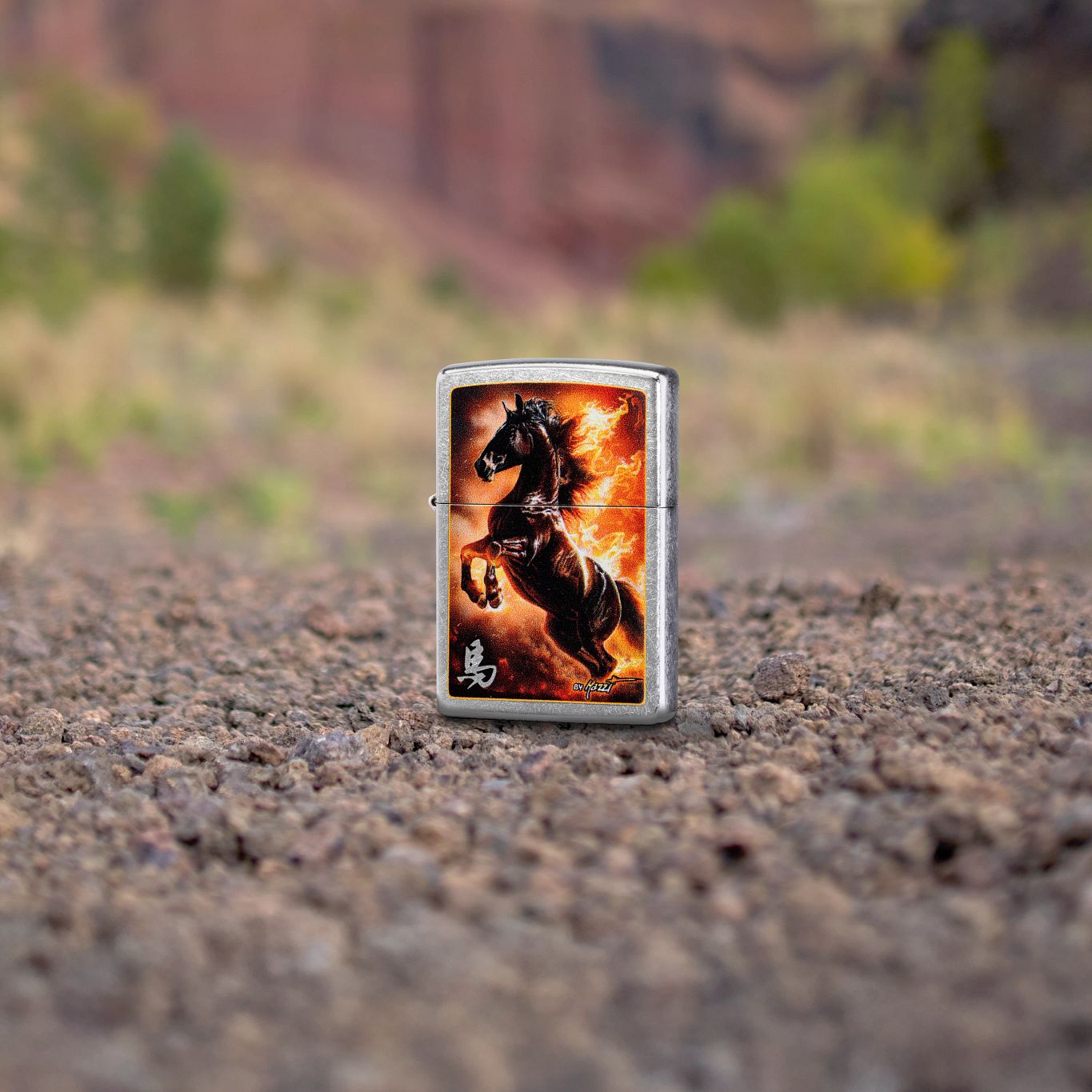 Lifestyle image of Zippo Mazzi® Fire Stallion Design Street Chrome Windproof Lighter standing in gravel with a nature background, showing the front of the lighter. 


