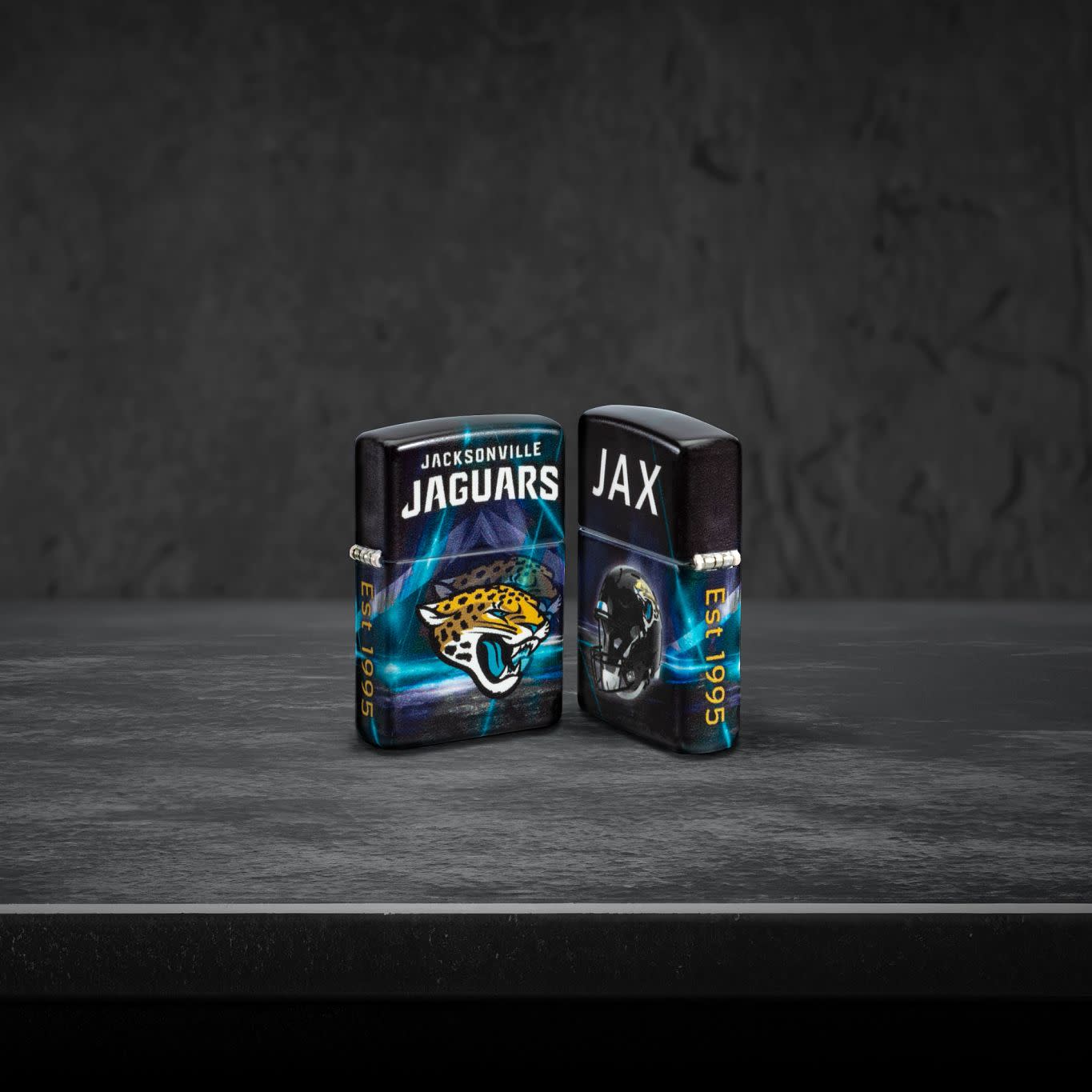 Lifestyle image of two Zippo NFL Jacksonville Jaguars 540 Matte Windproof Lighters, one showing the front of the lighter and the other showing the back, standing on a dark grey surface.