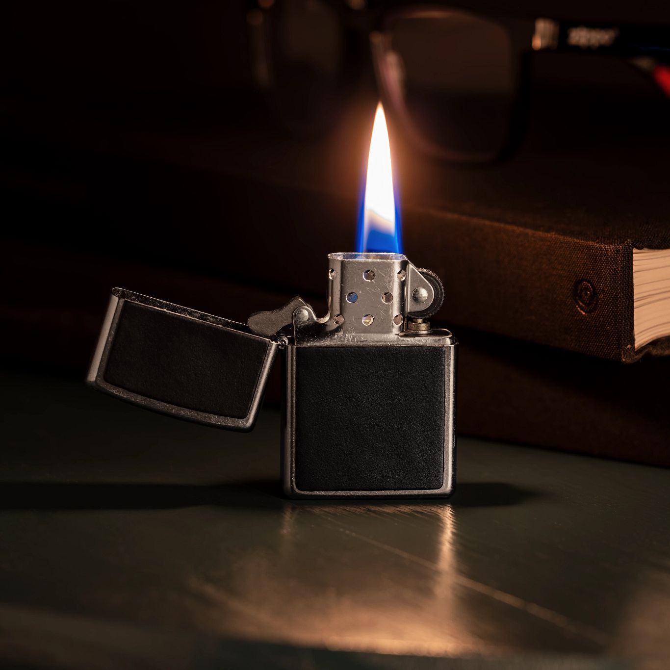 Lifestyle image of Zippo Black Leather Emblem Street Chrome Windproof Lighter standing on a desk, open and lit, next to books and a pair of eyeglasses. 

