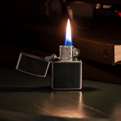 Lifestyle image of Zippo Black Leather Emblem Street Chrome Windproof Lighter standing on a desk, open and lit, next to books and a pair of eyeglasses. 

