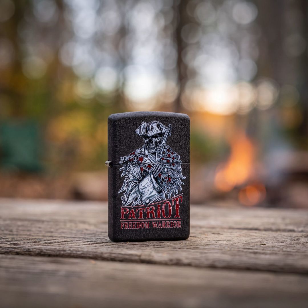 Lifestyle image of Zippo Freedom Warrior Design Black Crackle® Windproof Lighter standing on a weathered wooden surface showing the front and hinge side of the lighter with an out of focus campfire as the background. 

