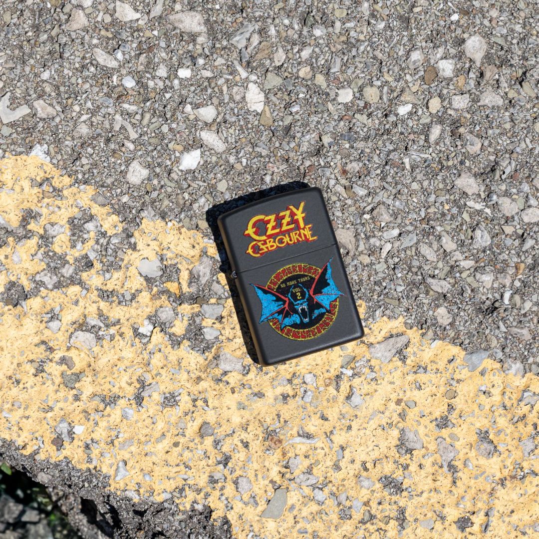 Lifestyle image of Zippo Ozzy Osbourne No More Tours Design Black Matte Windproof Lighter lying on the road showing the front of the lighter. 

