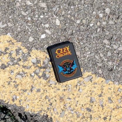 Lifestyle image of Zippo Ozzy Osbourne No More Tours Design Black Matte Windproof Lighter lying on the road showing the front of the lighter. 

