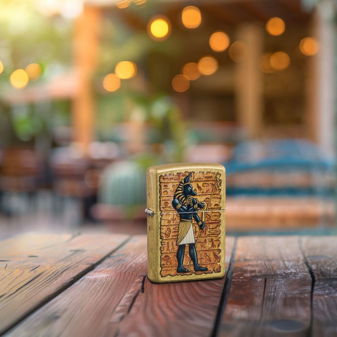 Lifestyle image of Zippo Guardian of Tombs Design Street Brass Windproof Lighter standing on a wooden surface showing the front and hinge side of the lighter with an out of focus restaurant as the background.

