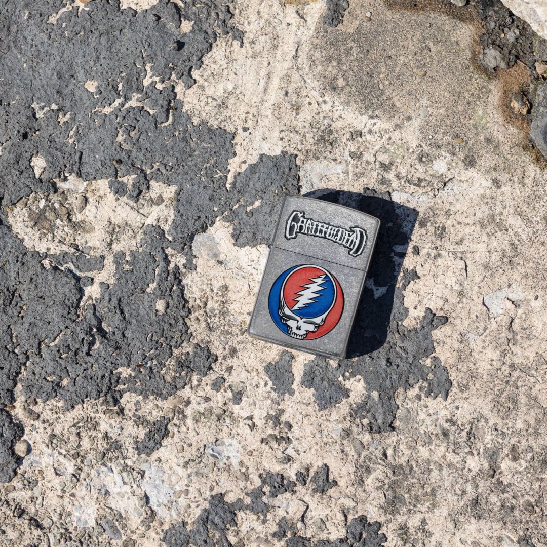 Lifestyle image of Zippo Grateful Dead Steal Your Face Design Street Chrome Windproof Lighter lying on weathered dark grey painted concrete, showing the front of the lighter. 

