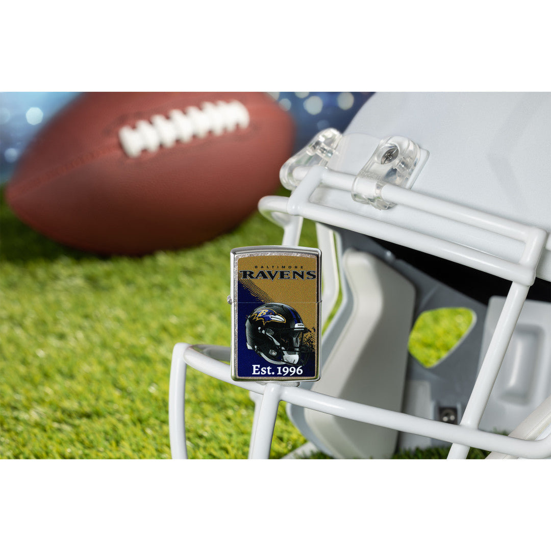 Lifestyle image of Zippo NFL Baltimore Ravens Helmet Design Street Chrome Windproof Lighter standing on the facemask of a white helmet showing the front of the lighter with a football lying on a football field as the background.

