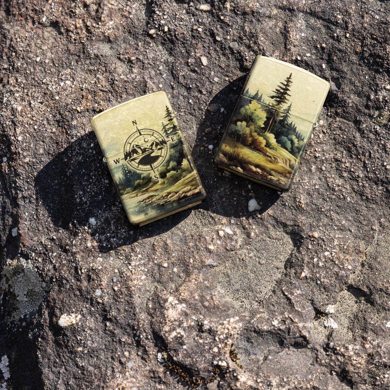 Lifestyle image of two Zippo Watercolor Wilderness Design 540 Tumbled Brass Windproof Lighters; one showing the front of the lighter and the other showing the back of the lighter, both lying on a boulder. 

