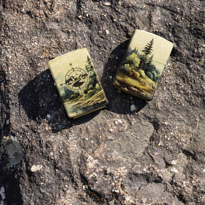 Lifestyle image of two Zippo Watercolor Wilderness Design 540 Tumbled Brass Windproof Lighters; one showing the front of the lighter and the other showing the back of the lighter, both lying on a boulder. 

