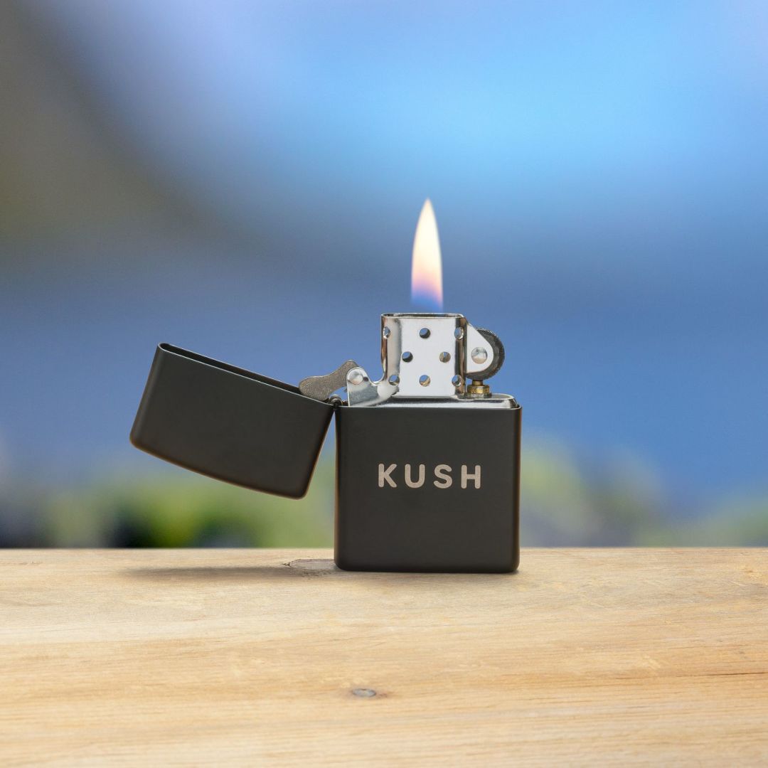 Lifestyle image of Zippo Kush Design Black Matte with Chrome Windproof Lighter standing on a wooden surface open and lit, showing the front of the lighter with a few treetops and the blue sky as the background.

