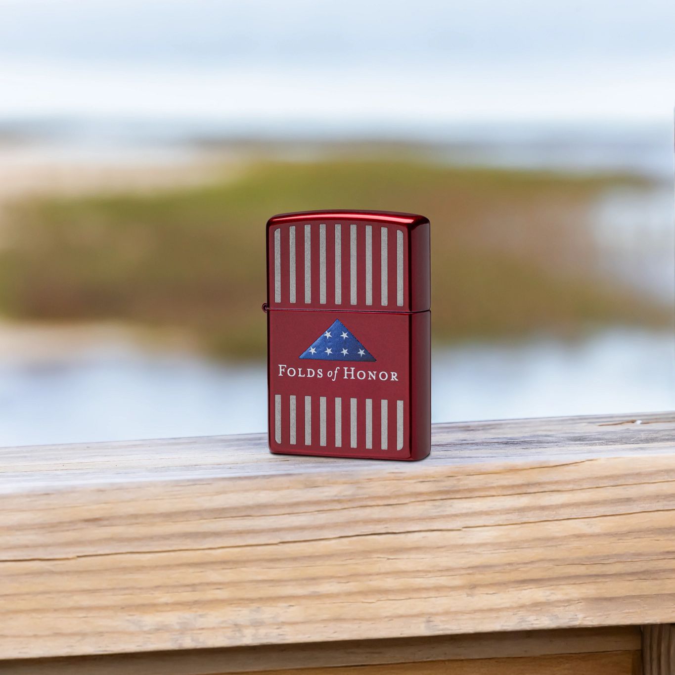 Lifestyle image of Zippo Folds of Honor Pinstripes Design Candy Apple Red Windproof Lighter standing on a wooden deck railing with a grassy body of water as the background, showing the front of the lighter. 

