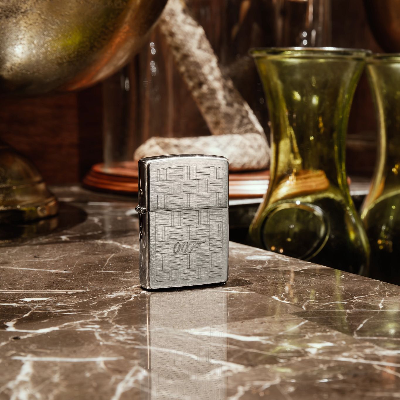 Lifestyle image of Zippo James Bond® Tuxedo Design Brushed Chrome Windproof Lighter standing on a brown and white stone countertop with green glass vases and other decor in the background.

