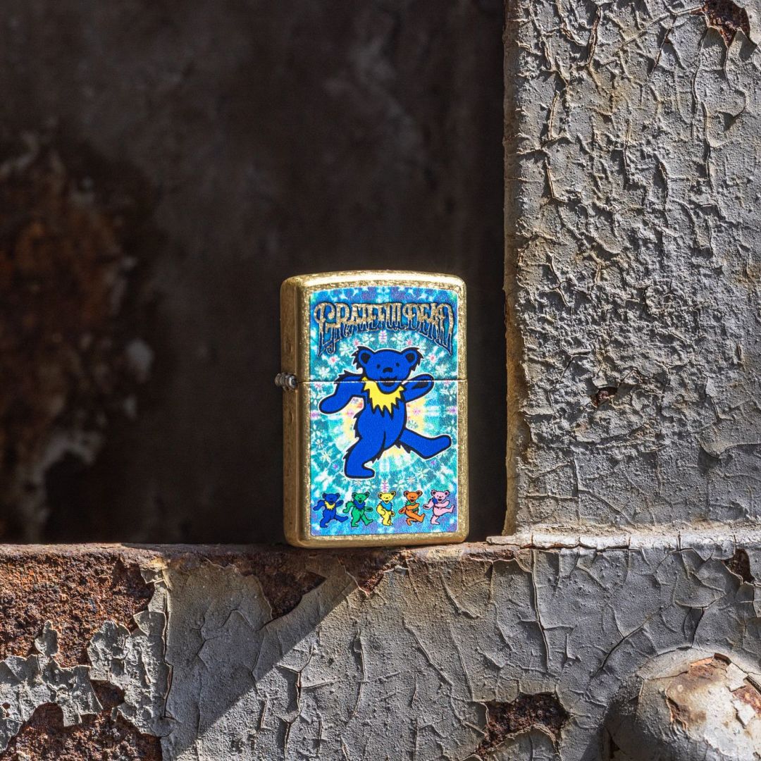 Lifestyle image of Zippo Grateful Dead Dancing Bears Design Street Brass Windproof Lighter standing on a rusty painted steel structure showing the front of the lighter. 

