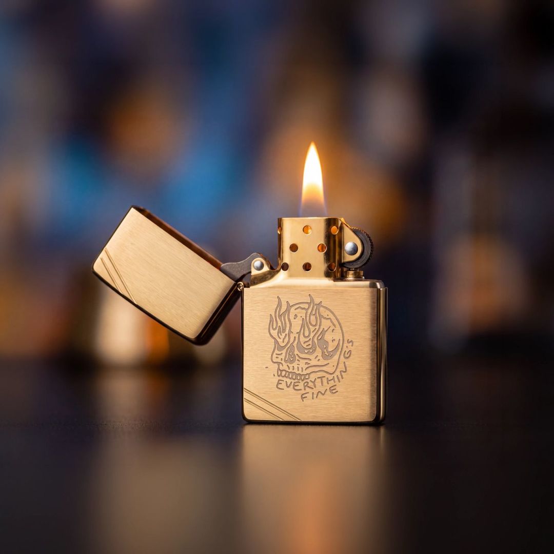 Lifestyle image of Zippo Everything's Fine Design Vintage Brushed Brass Windproof Lighter standing open and lit showing the front of the lighter with a dark out of focus background. 

