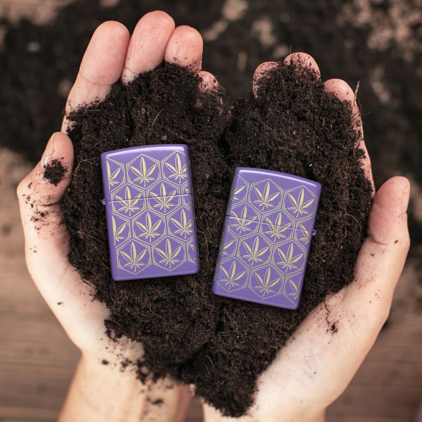 Lifestyle image of two Zippo Honeycomb Cannabis Design Smoky Lavender Windproof Lighters; one showing the front of the lighter and the other showing the back of the lighter, both being held while lying on top of soil with a blurred garden work area background. 

